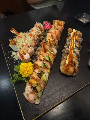 Assorted sushi rolls on a black plate, featuring shrimp tempura, spicy tuna, and eel rolls with colorful garnishes.
