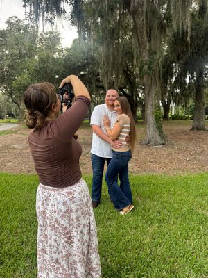 A photographer captures a couple embracing and smiling outdoors under Spanish moss-draped oak trees on a green lawn.
