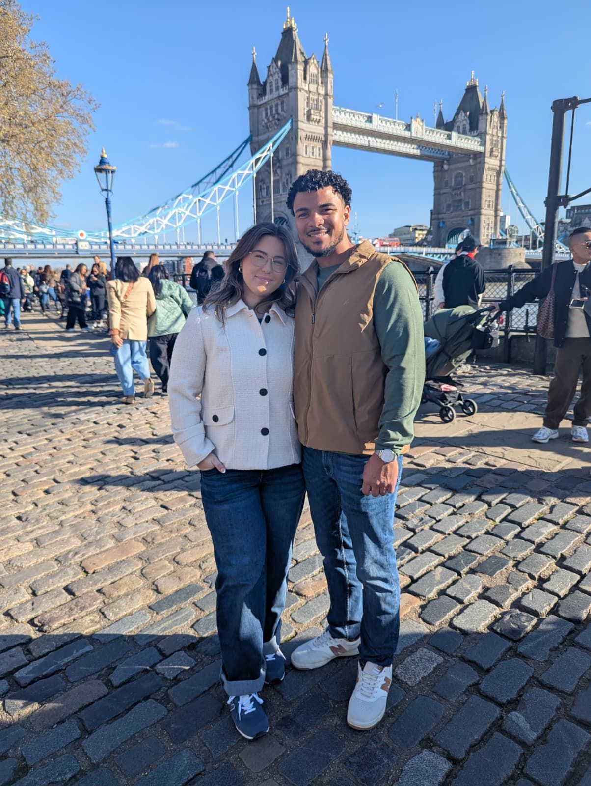 Couple posing in front of Tower Bridge in London on a sunny day with tourists in the background.