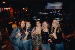 Five women pose together making rock signs at a lively bar venue with stage lighting in background.