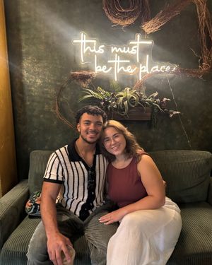 Couple smiling together on a green couch beneath a neon sign reading this must be the place.