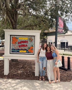 Three women pose together in front of a colorful mosaic Mount Dora welcome sign surrounded by trees.