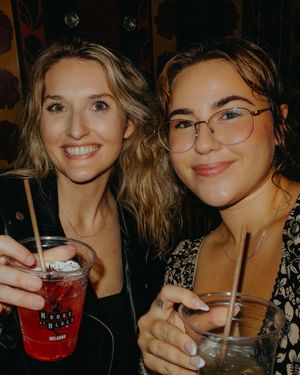 Two smiling women holding cocktails at House of Blues, one wearing glasses and a floral top.