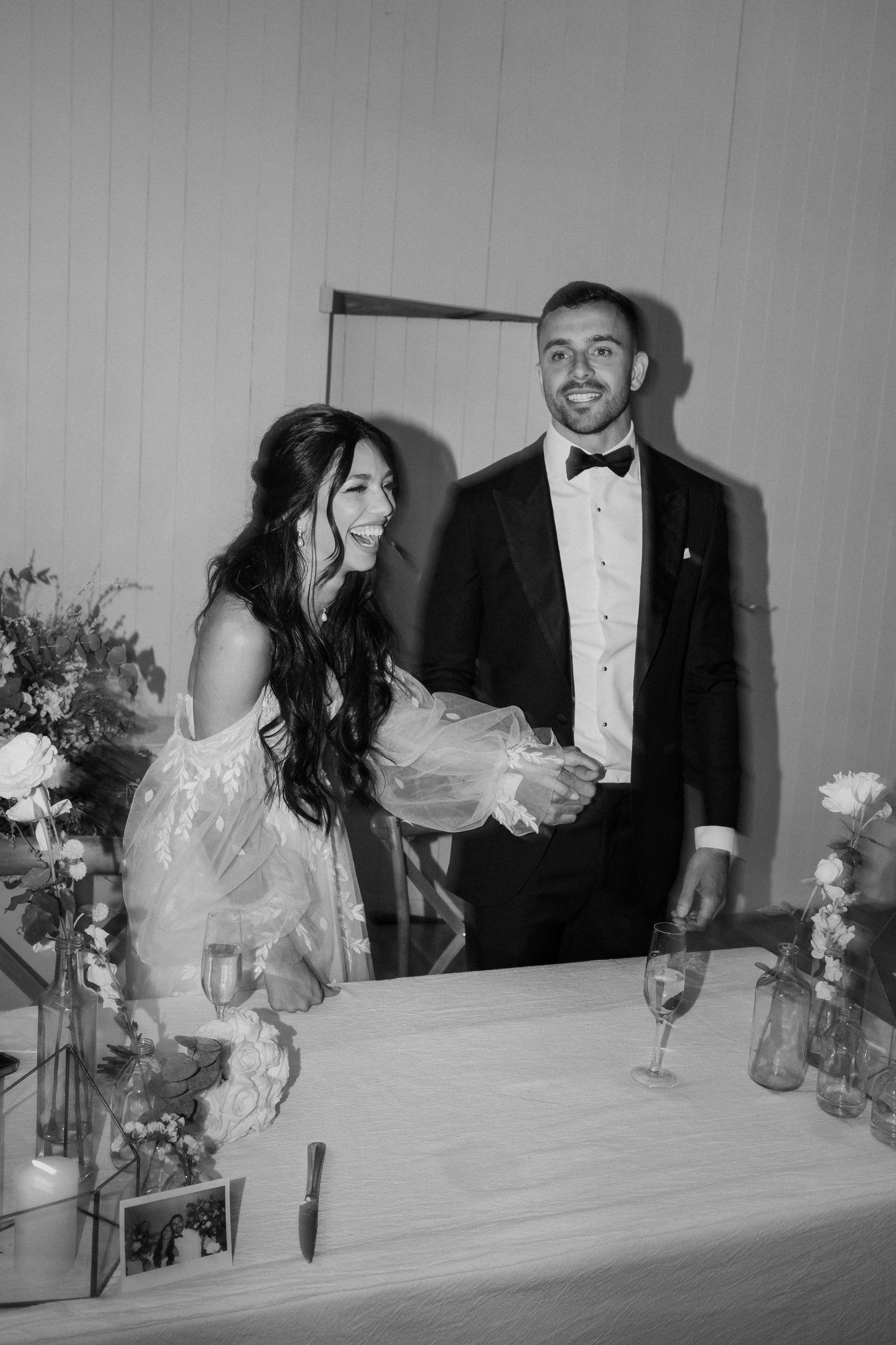Black and white photo of a laughing bride and smiling groom in tuxedo standing at a decorated wedding reception table.