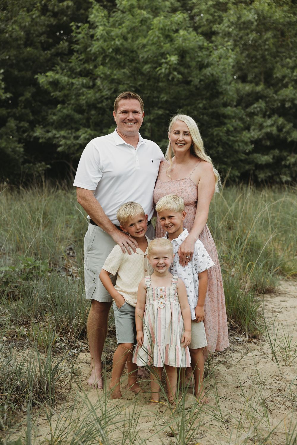 Happy family of five posing outdoors on sandy path surrounded by green trees and grass.