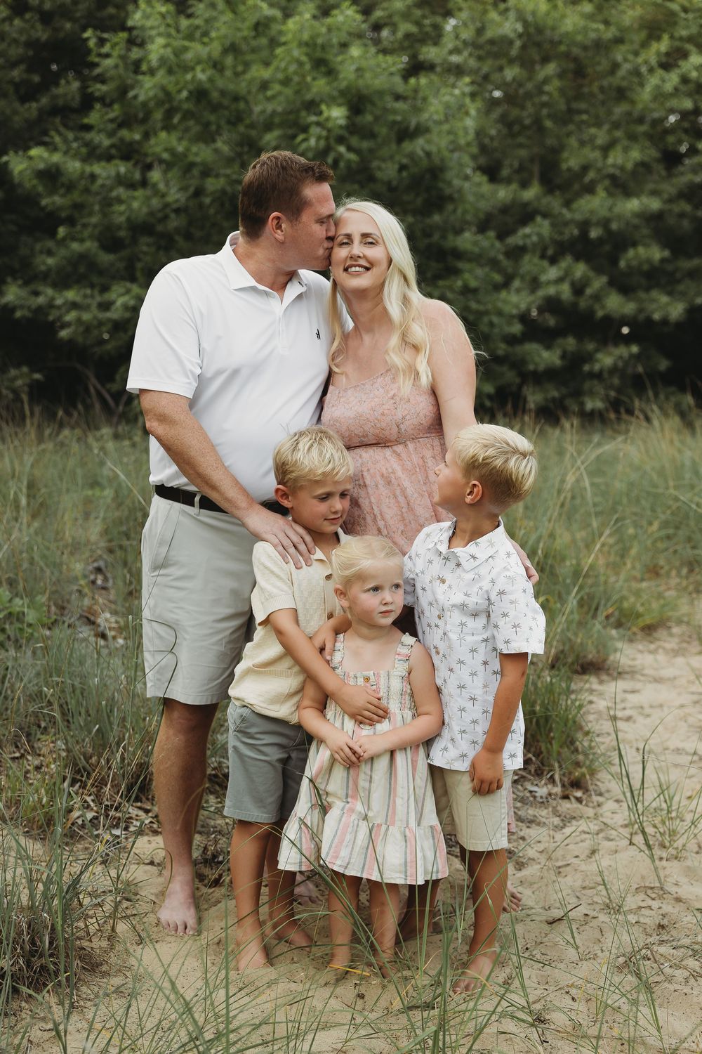 Family of five posing outdoors on sandy ground with lush green trees, parents with three young blonde children.