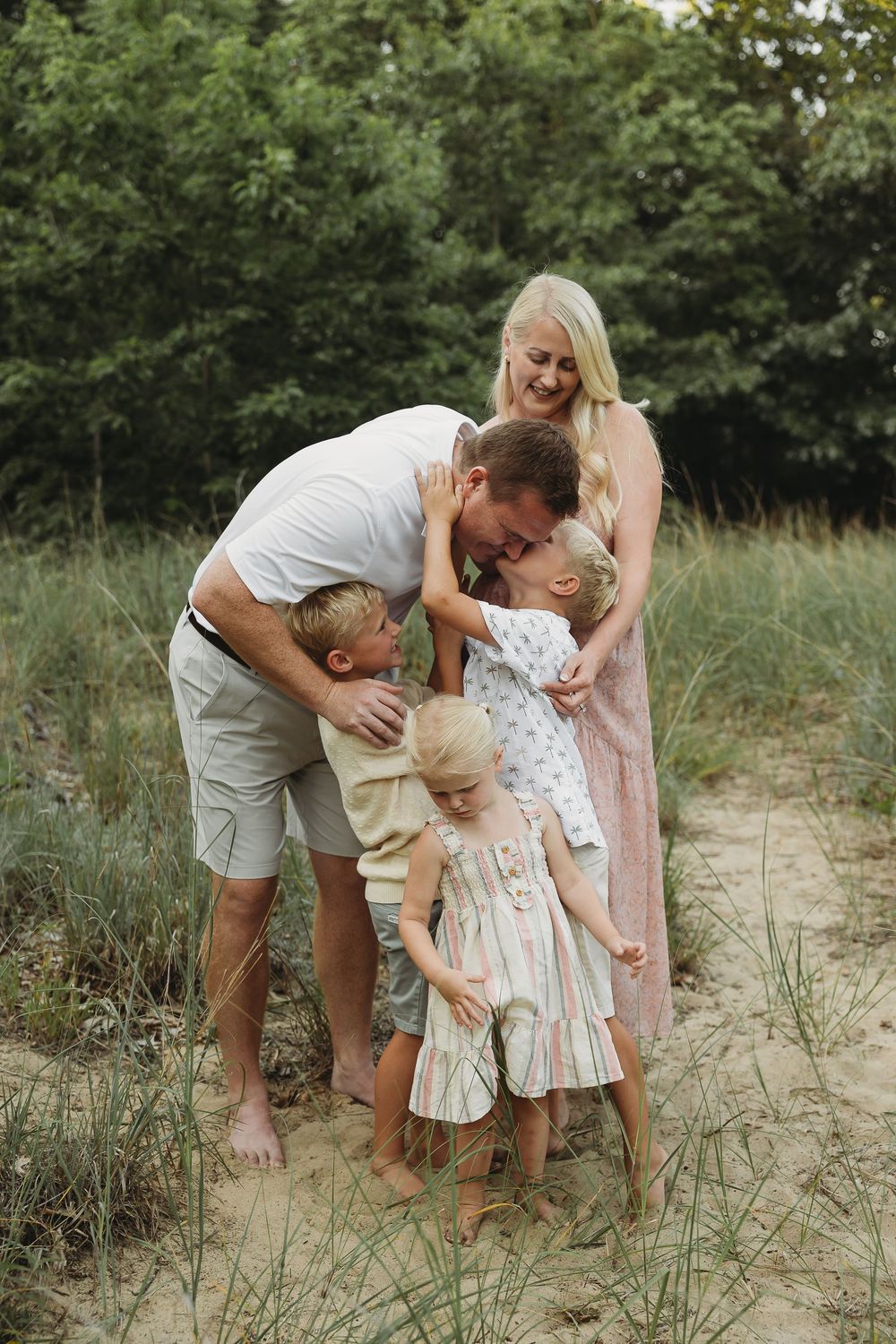 A happy family of five sharing a warm embrace on a sandy path surrounded by lush green trees.