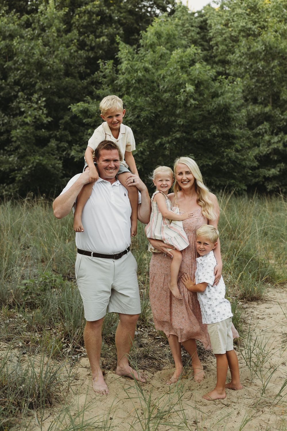 A smiling family of five poses outdoors on sandy ground with lush green trees in the background.