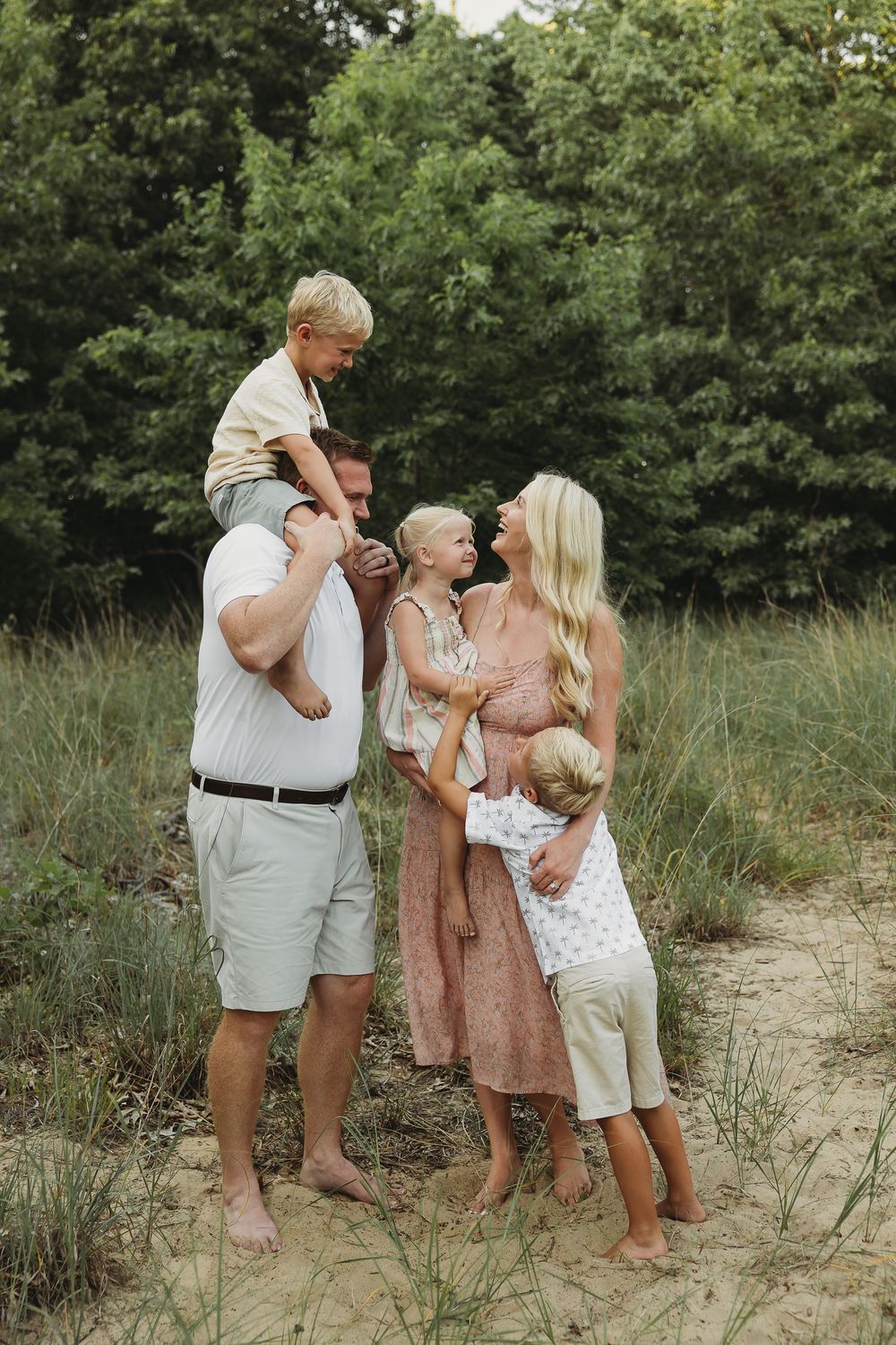 Happy family of five enjoying a sunny outdoor photo session in a sandy clearing surrounded by lush green trees.