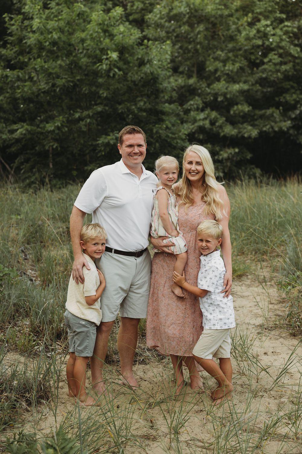 Happy family of five posing on sandy beach path surrounded by lush green trees on a summer day.