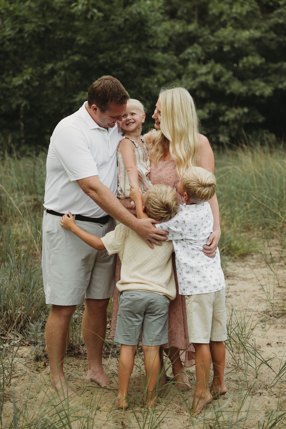 A happy family of five shares a warm embrace on a sandy beach surrounded by lush green trees.