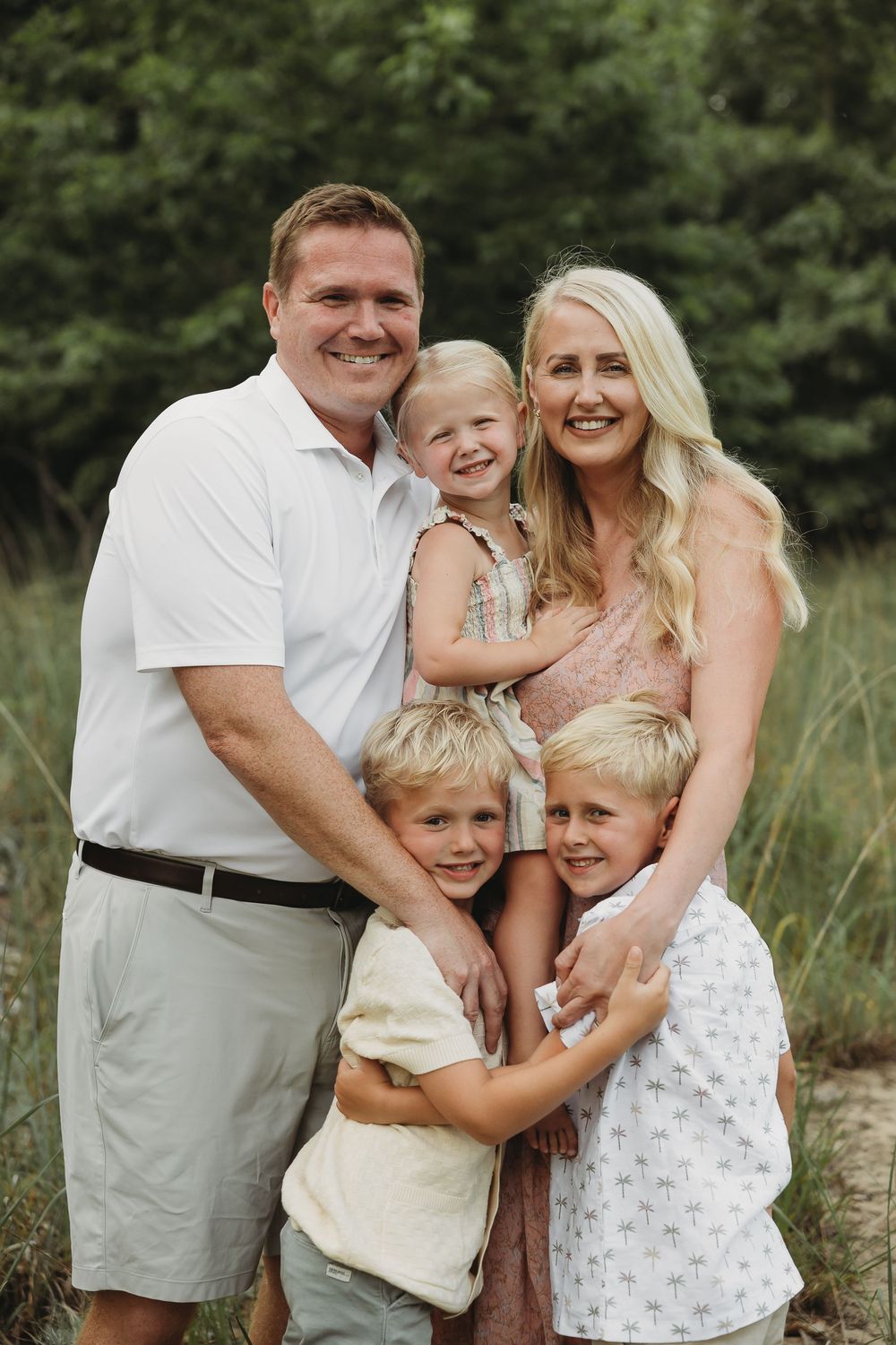 Happy family of five posing outdoors with lush green trees, parents smiling with three young blonde children.