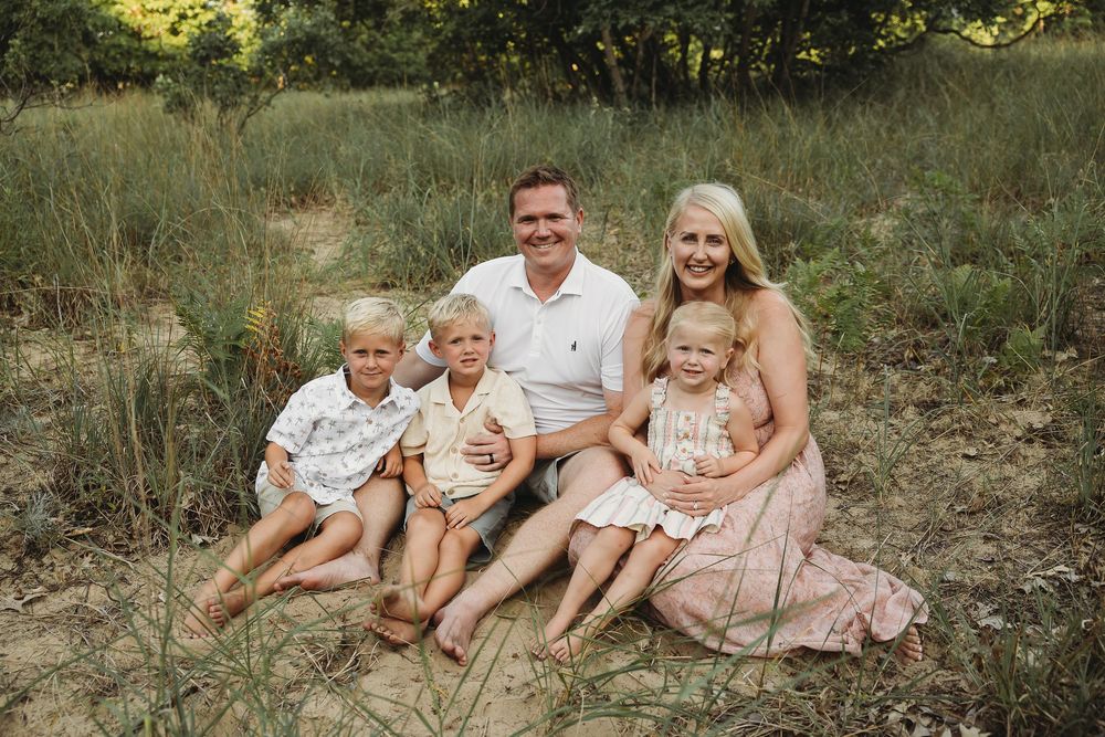 Family of five smiling outdoors in a grassy field, parents with three young children sitting together for a portrait.