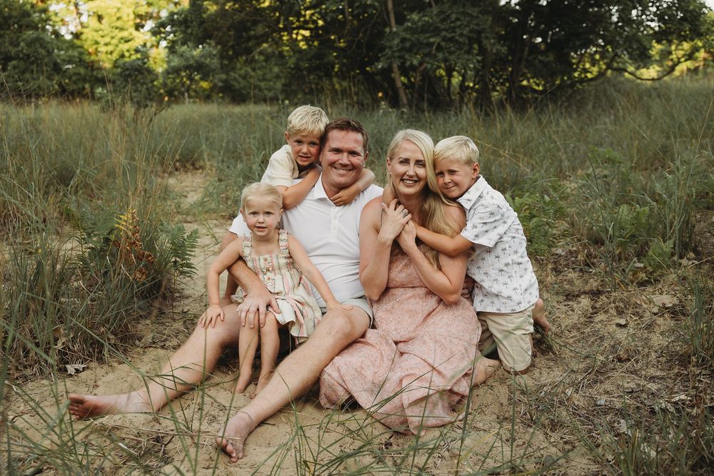 Happy family of five posing outdoors on sandy ground surrounded by lush green trees and grass.