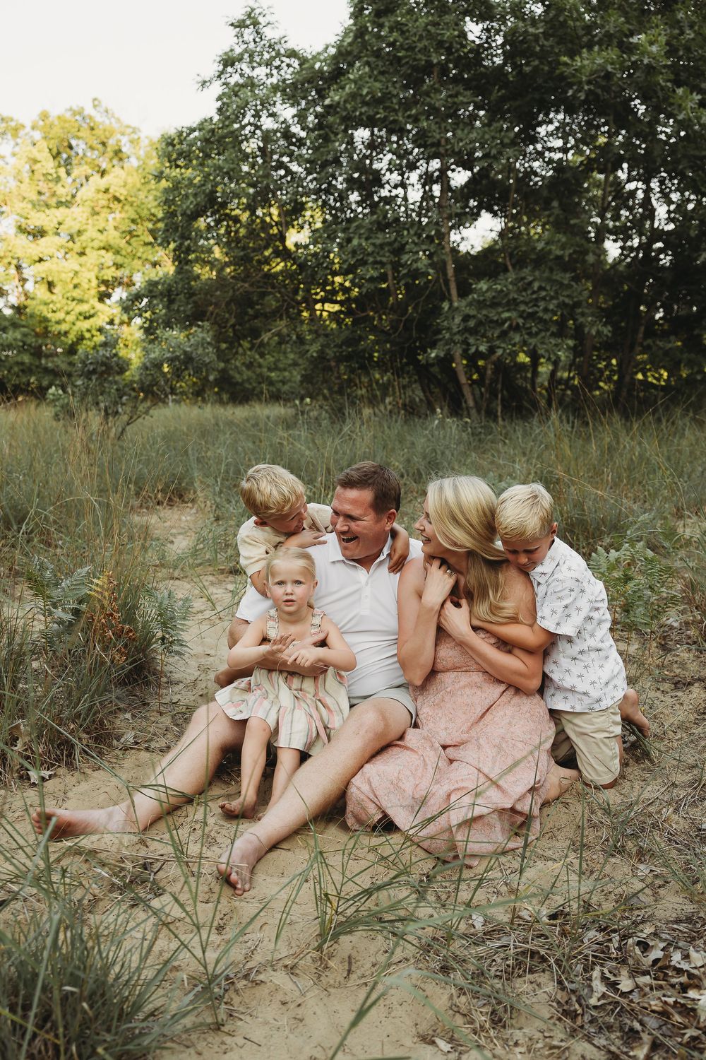 A joyful family of five sits together on sandy ground surrounded by lush green trees and tall grass outdoors.