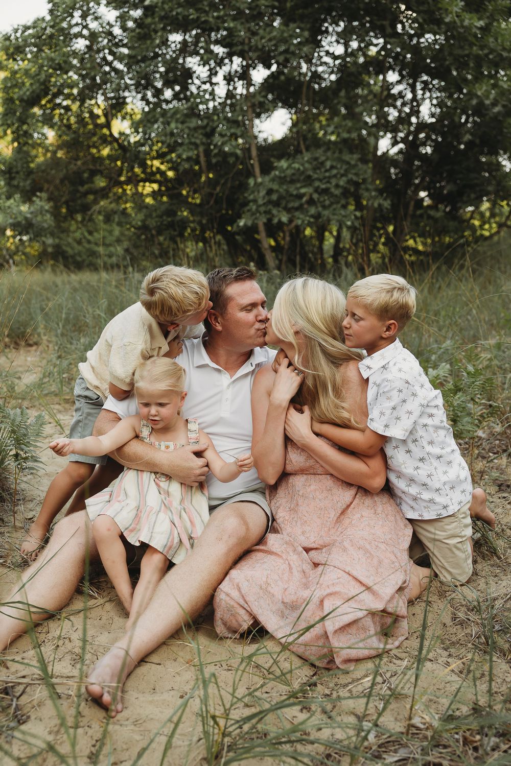 Family of five sitting together on sandy ground surrounded by lush green trees, parents and three blonde children sharing a warm embrace.