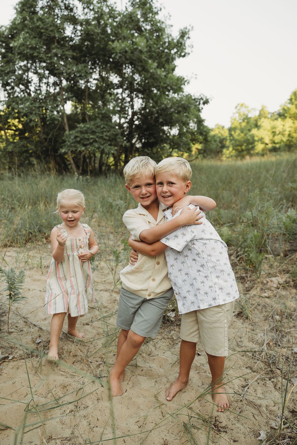 Two blonde boys hugging on sandy beach path with toddler girl in pink dress, trees in background.