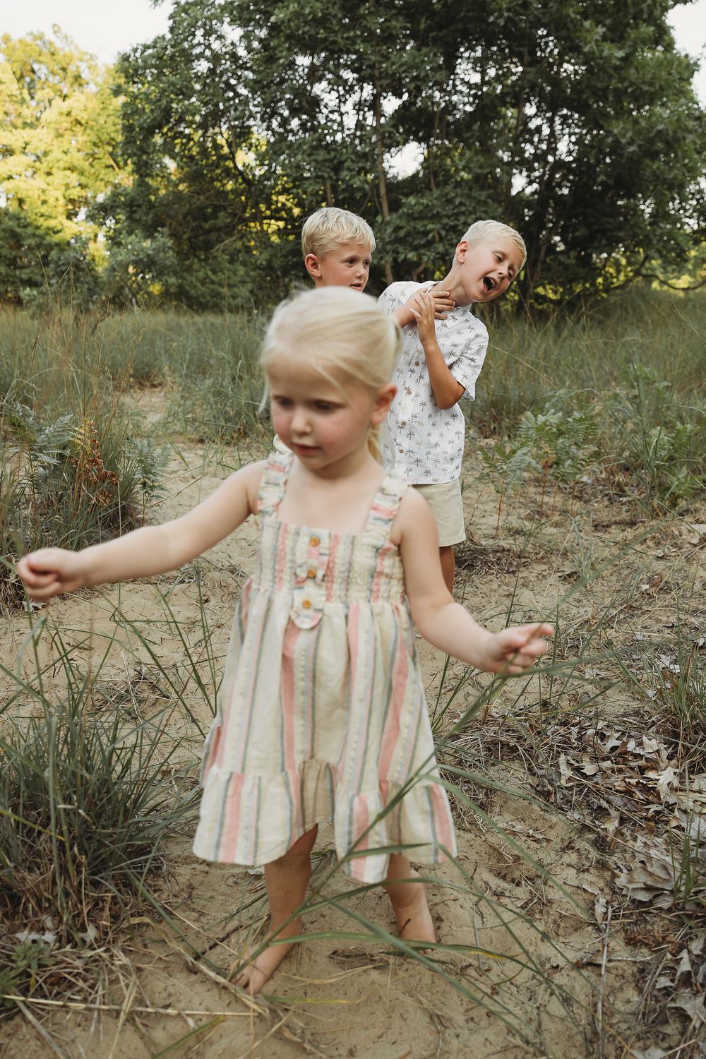 Three young blonde children playing outdoors in a sandy woodland area, with a girl in a striped dress in the foreground.