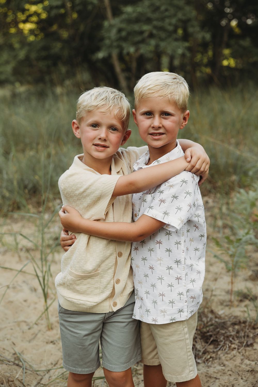 Two blonde boys hugging each other outdoors on sandy ground with green trees in the background.