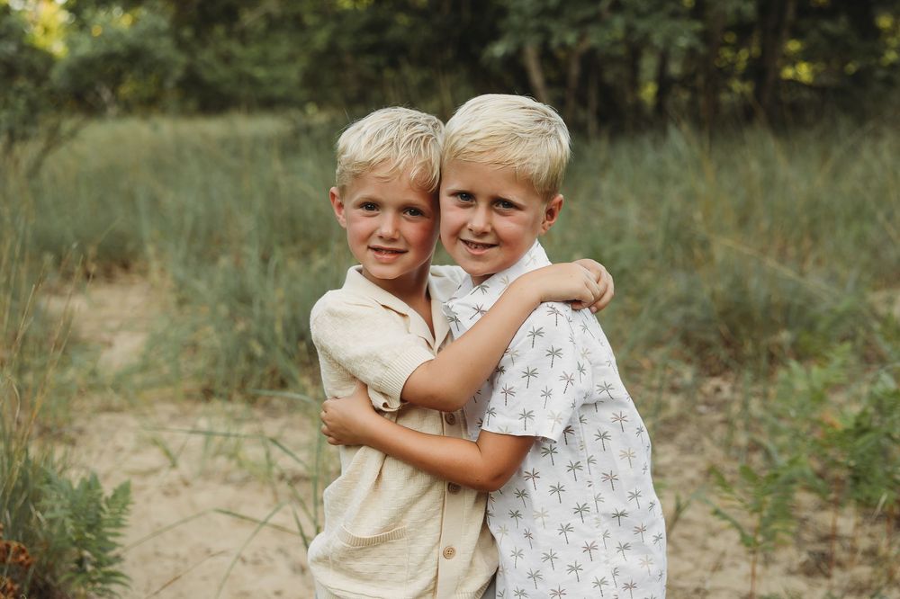 Two blonde boys hugging and smiling outdoors on sandy path with green trees in background.