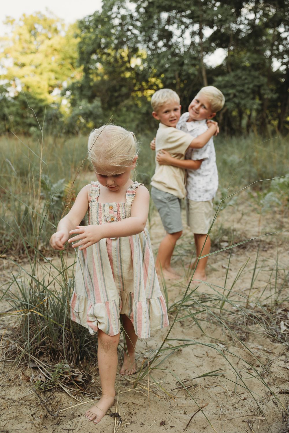 Three blonde children playing outdoors in nature, two boys hugging while a young girl in striped dress explores sandy ground.