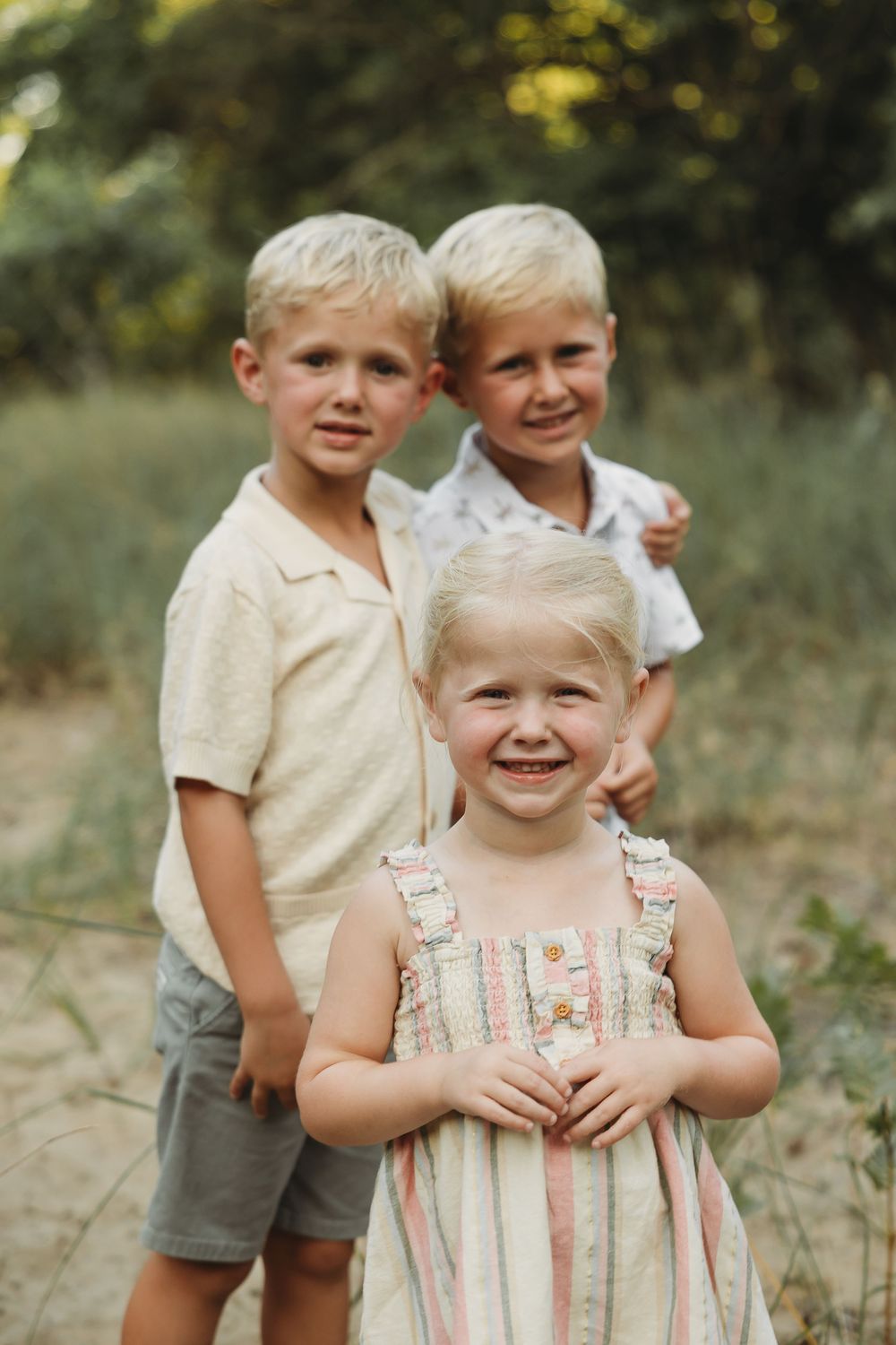 Three blonde children pose outdoors, two boys standing behind a smiling toddler girl in a striped dress.