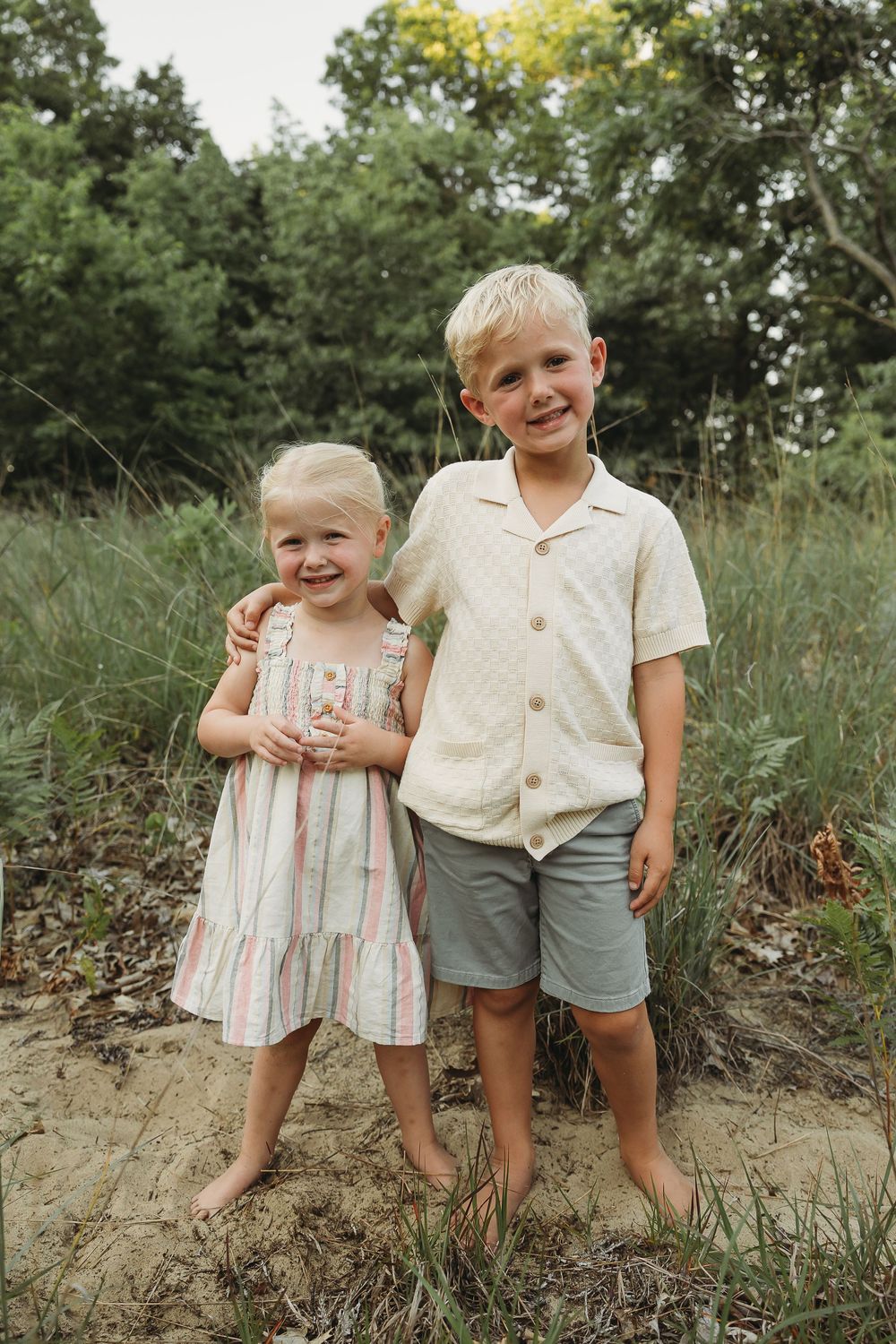 Two young blonde siblings pose together outdoors, girl in striped dress and boy in cream shirt with gray shorts.