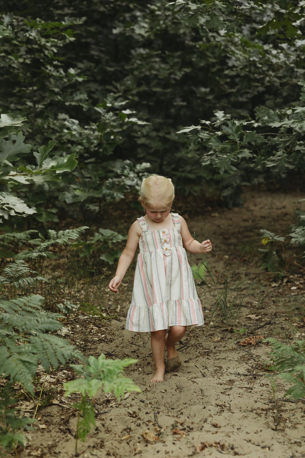 Young blonde toddler girl in striped dress walking barefoot on a dirt path surrounded by lush green forest foliage.