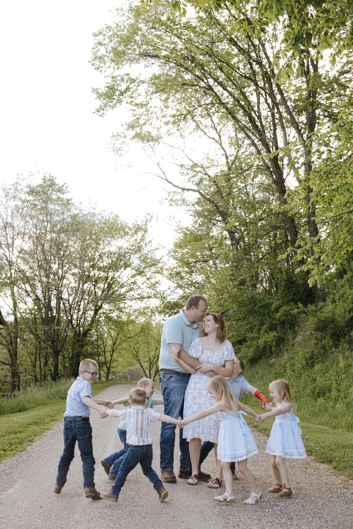 Family of six poses on a tree-lined path, parents kissing while four young children playfully hold hands around them.