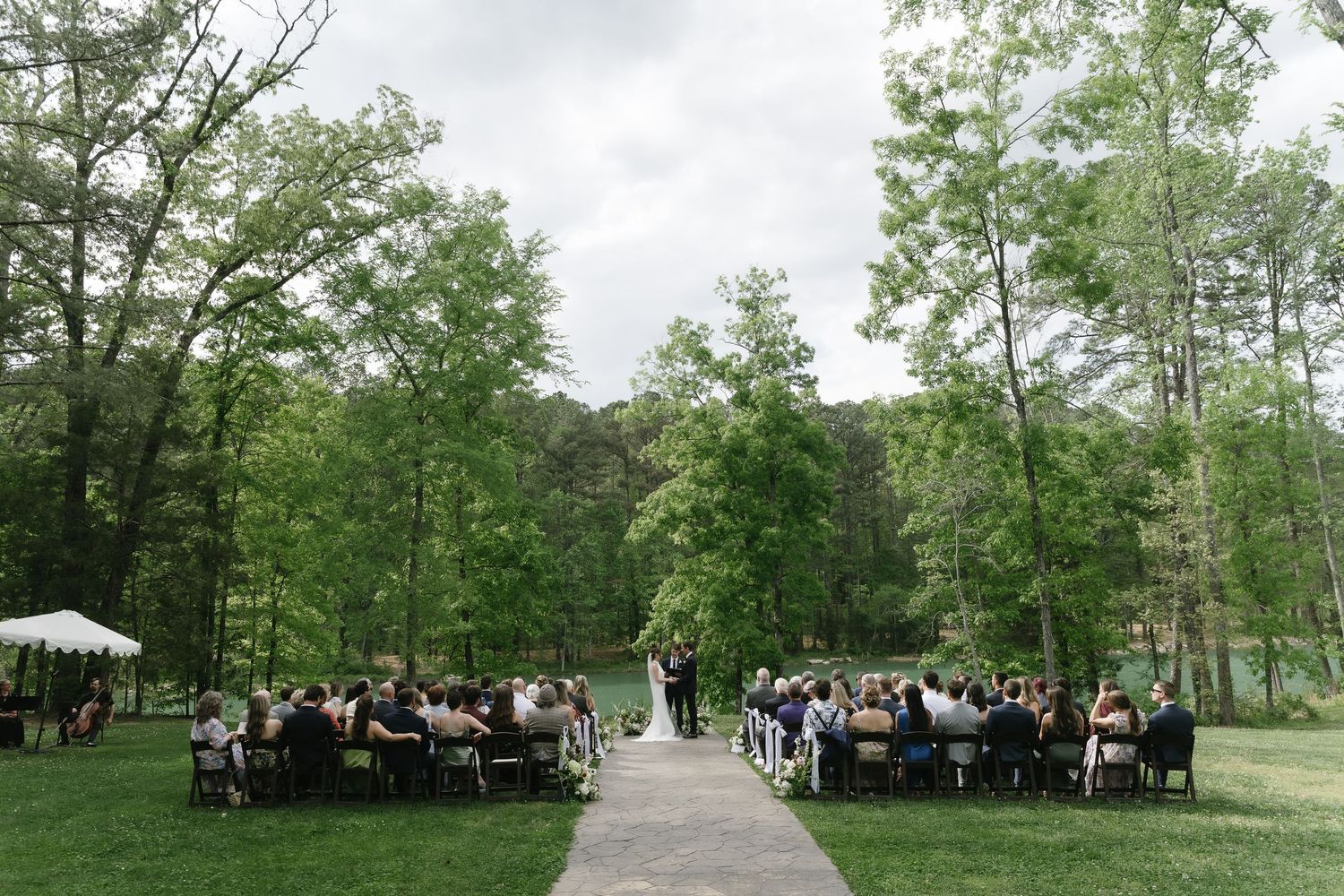 Outdoor wedding ceremony on a lush green lawn surrounded by tall trees with guests seated in rows.