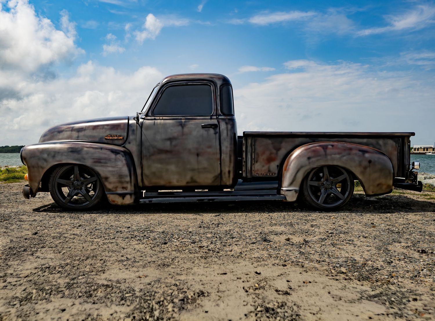 Vintage patina rat rod pickup truck parked on gravel near water under a blue sky with clouds.