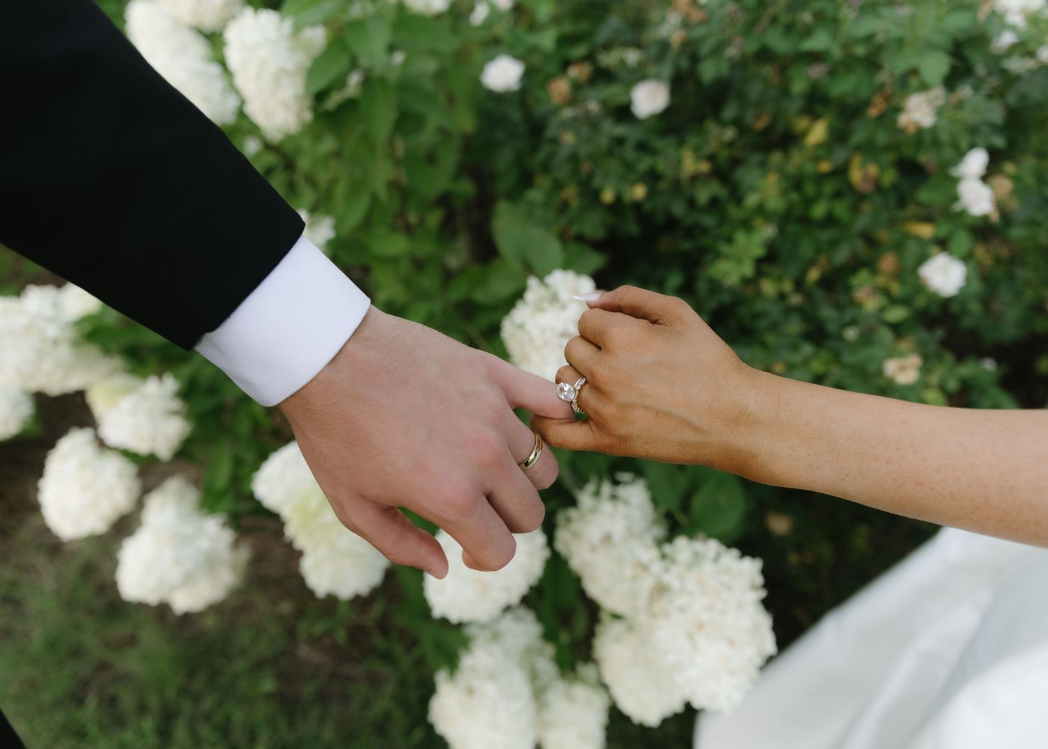 Bride and groom holding hands with wedding rings, white hydrangeas in background.