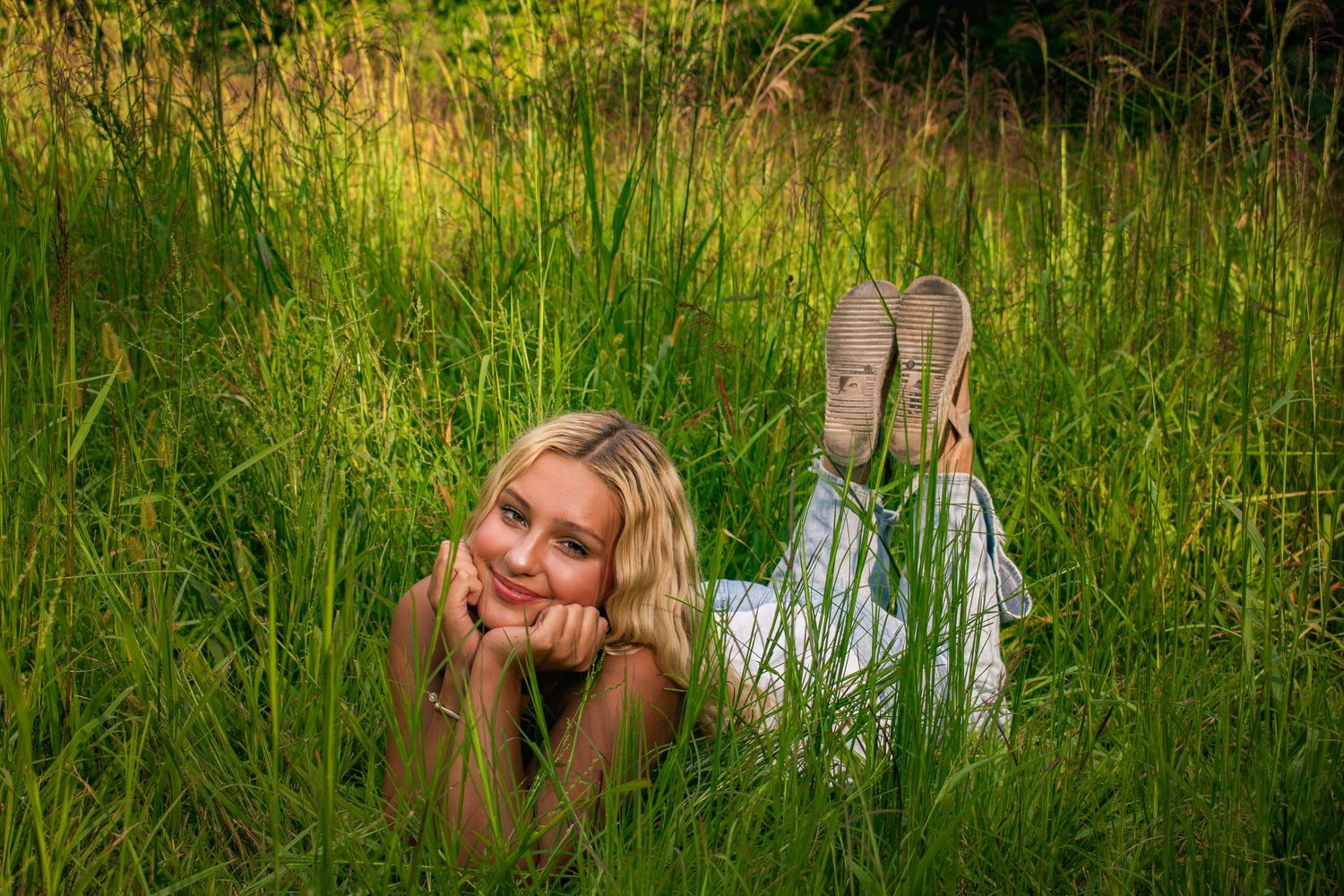 Smiling blonde girl lying in lush tall green grass wearing white outfit with shoes visible.
