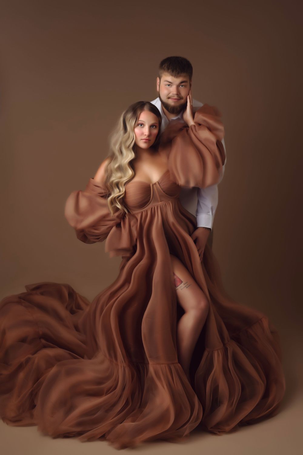 Pregnant woman in flowing brown gown poses with man in studio maternity portrait photography.
