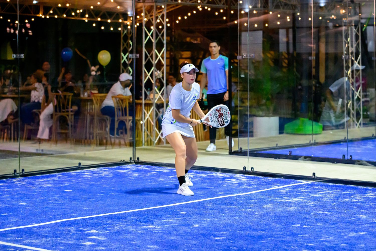 Female padel tennis player in white outfit mid-swing on blue court with audience and festive lights behind.