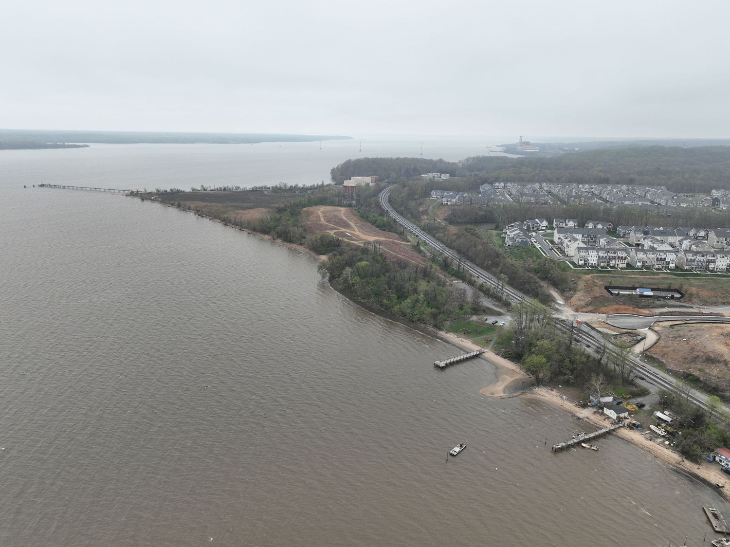 Aerial view of a coastal waterway with sandy shores, dense trees, and a residential neighborhood under overcast skies.