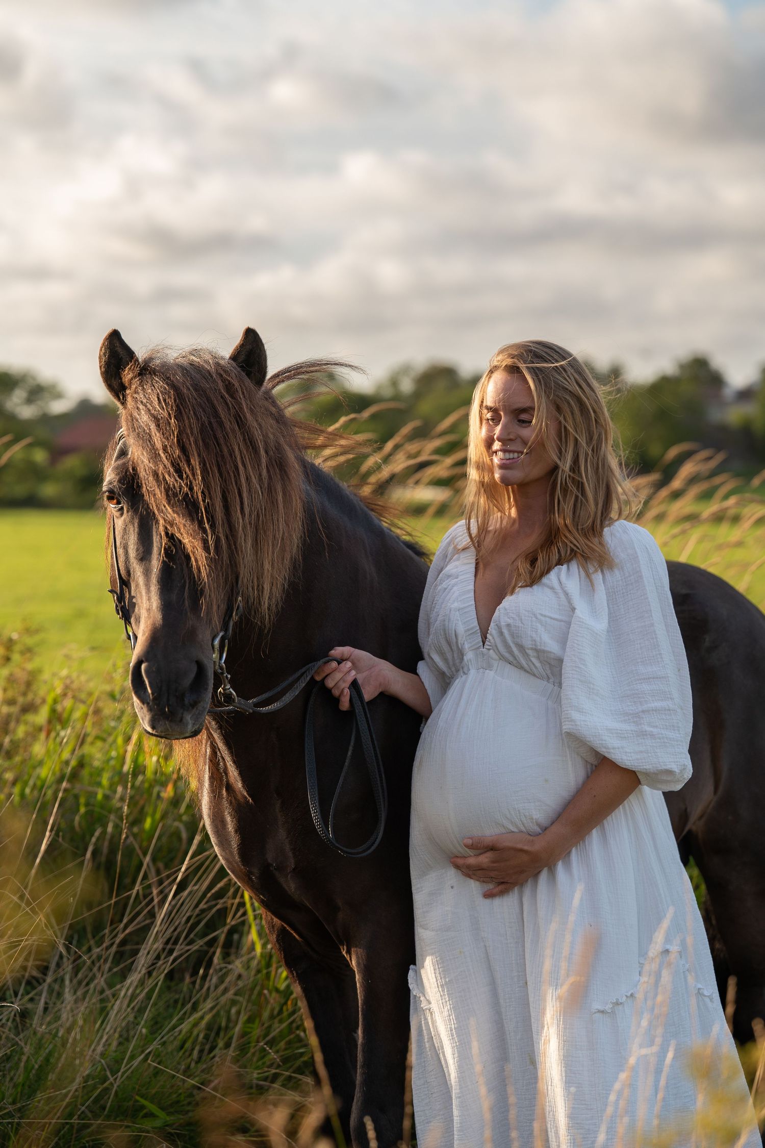 Smiling pregnant woman in white dress holding dark horse lead.