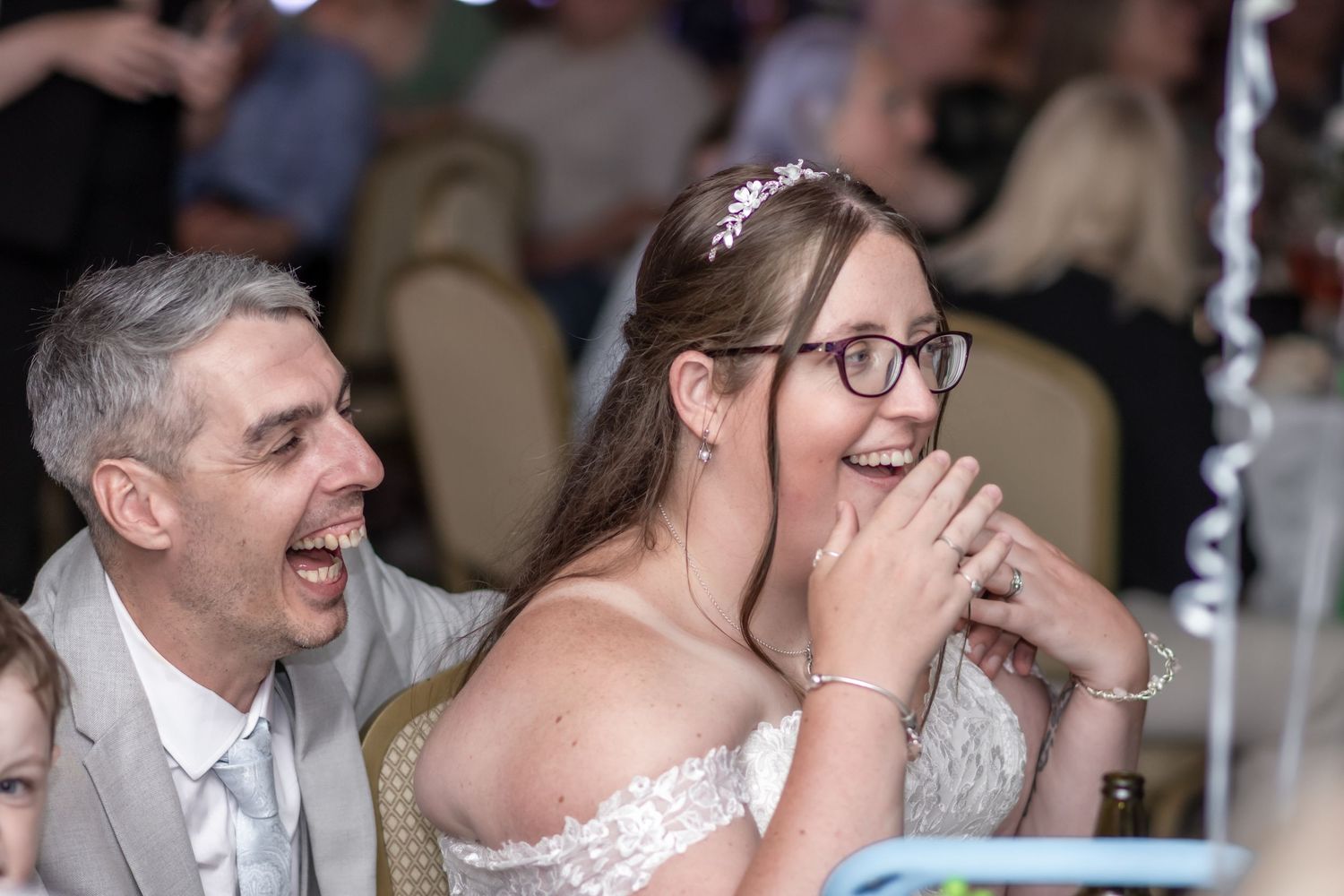 Bride and groom laughing joyfully together at their wedding reception, bride wearing white lace dress and glasses.