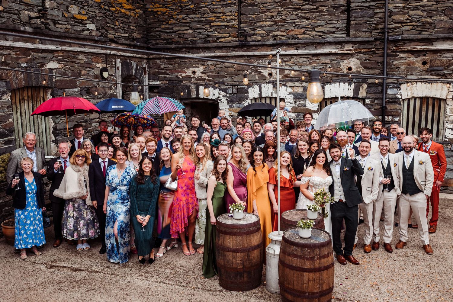 Large wedding group photo outdoors at rustic stone venue with colorful attire and umbrellas on a rainy day.