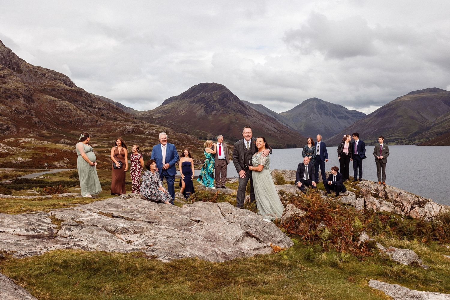 Large wedding party group posing outdoors in formal attire on green lawn with garden backdrop.