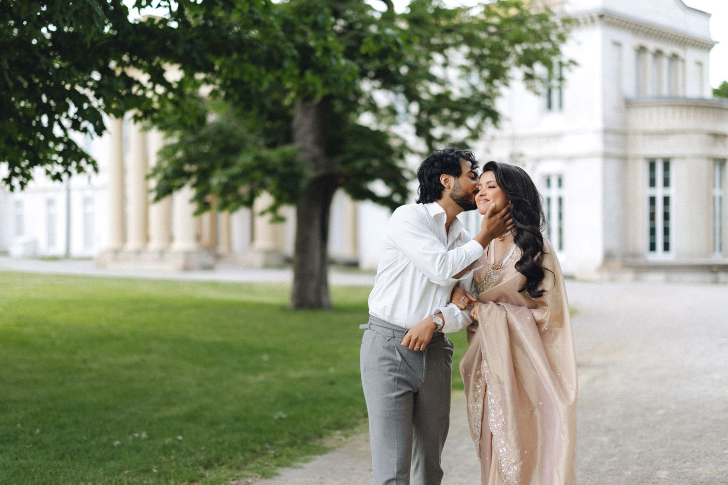 Couple relaxing on green grass in a park, woman in pink dress lying on man's lap near a white building.