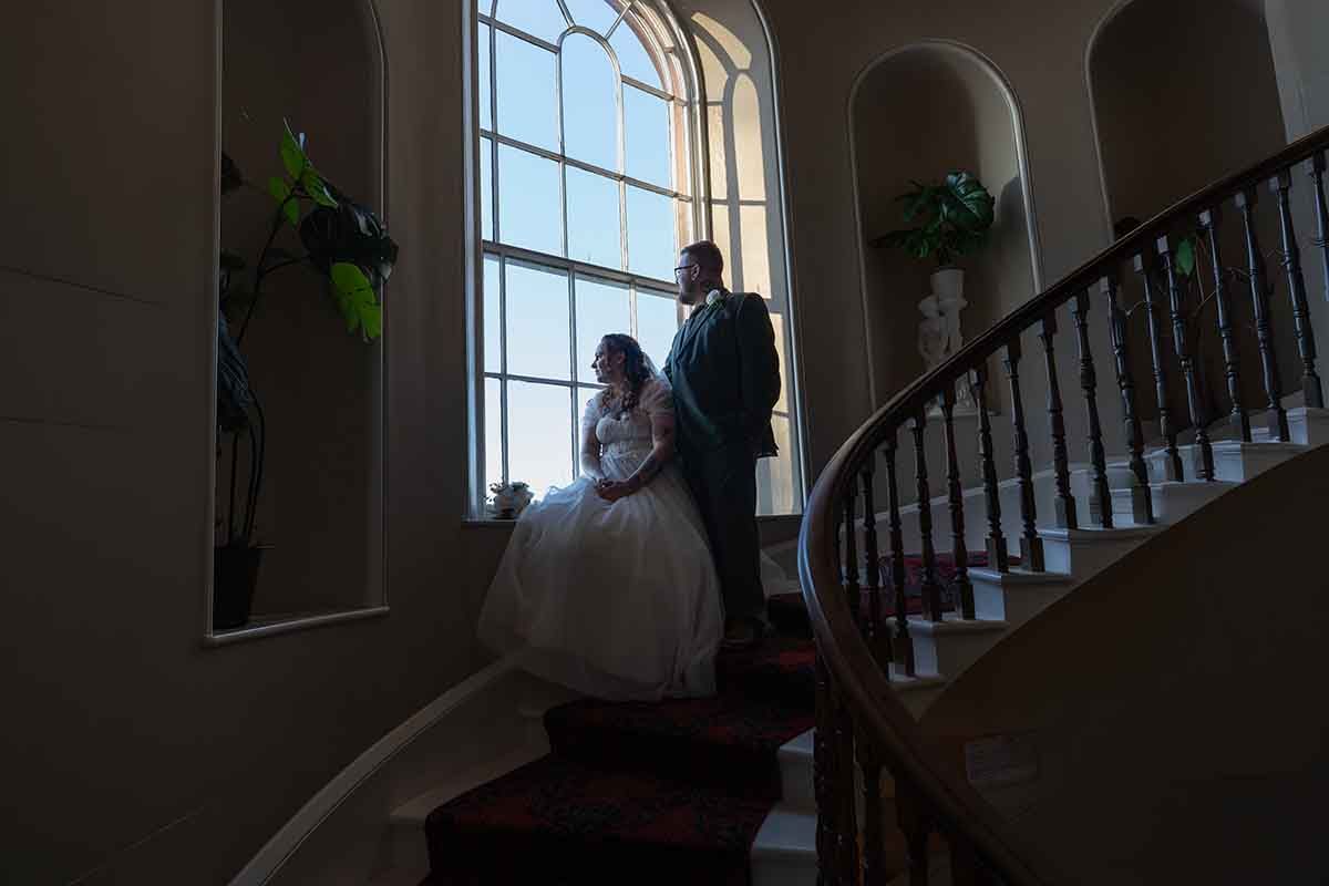 Bride and groom stand by a grand arched window on an elegant staircase in a dimly lit wedding venue.