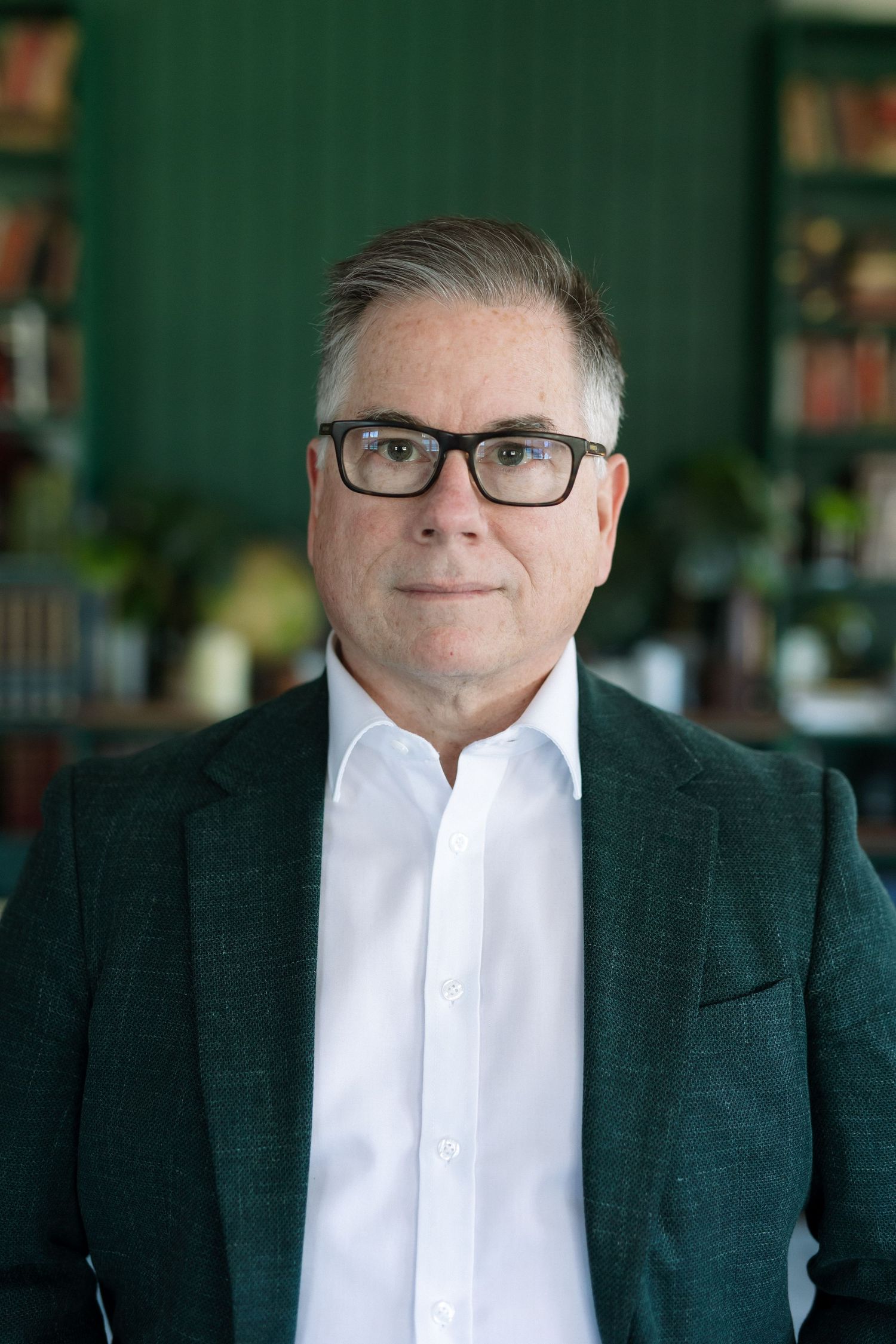 Middle-aged man with gray hair and black glasses wearing a dark green blazer and white shirt in a library setting.