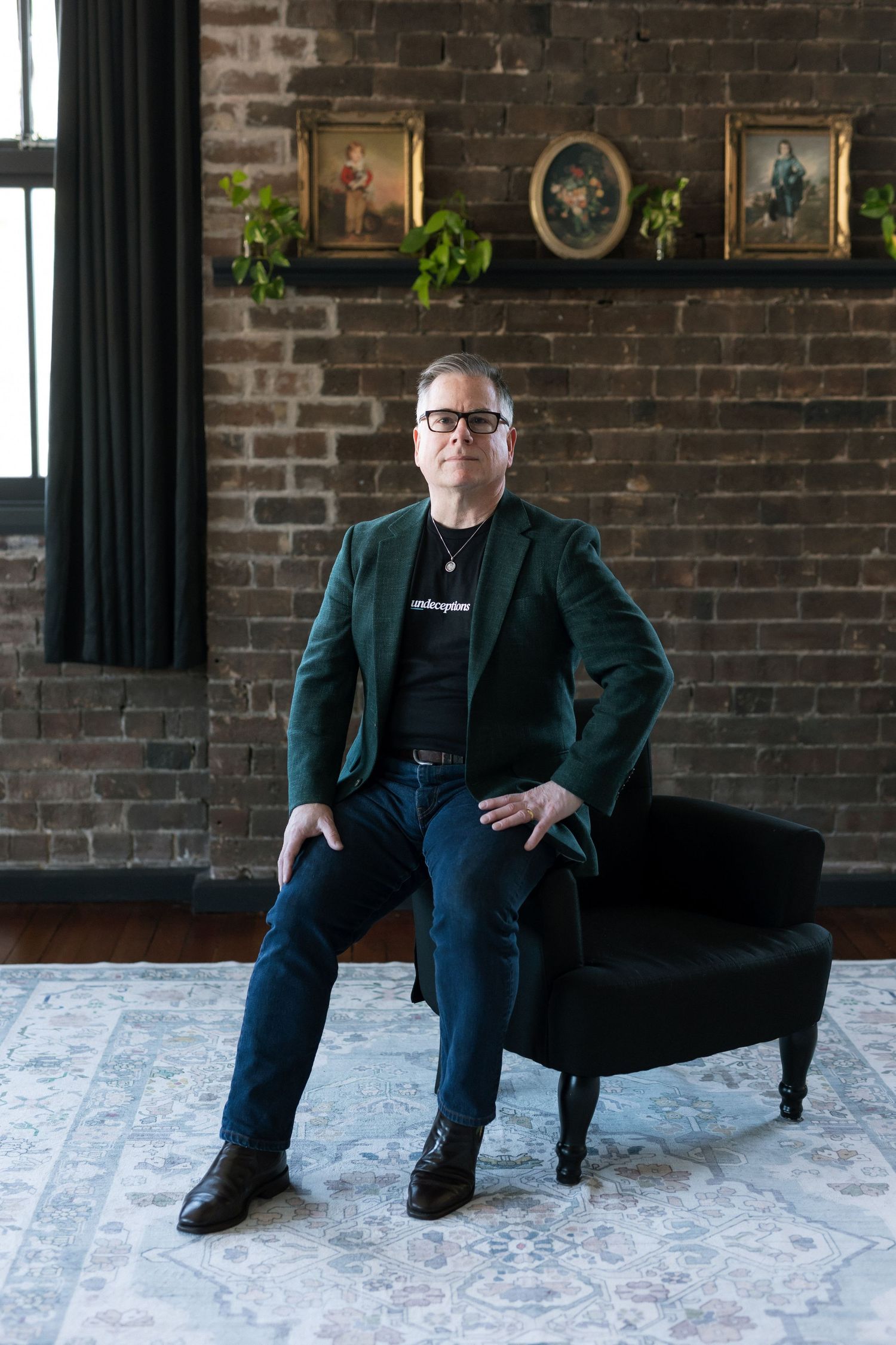 Man in green blazer and jeans sits on black chair against brick wall with framed portraits and plants.