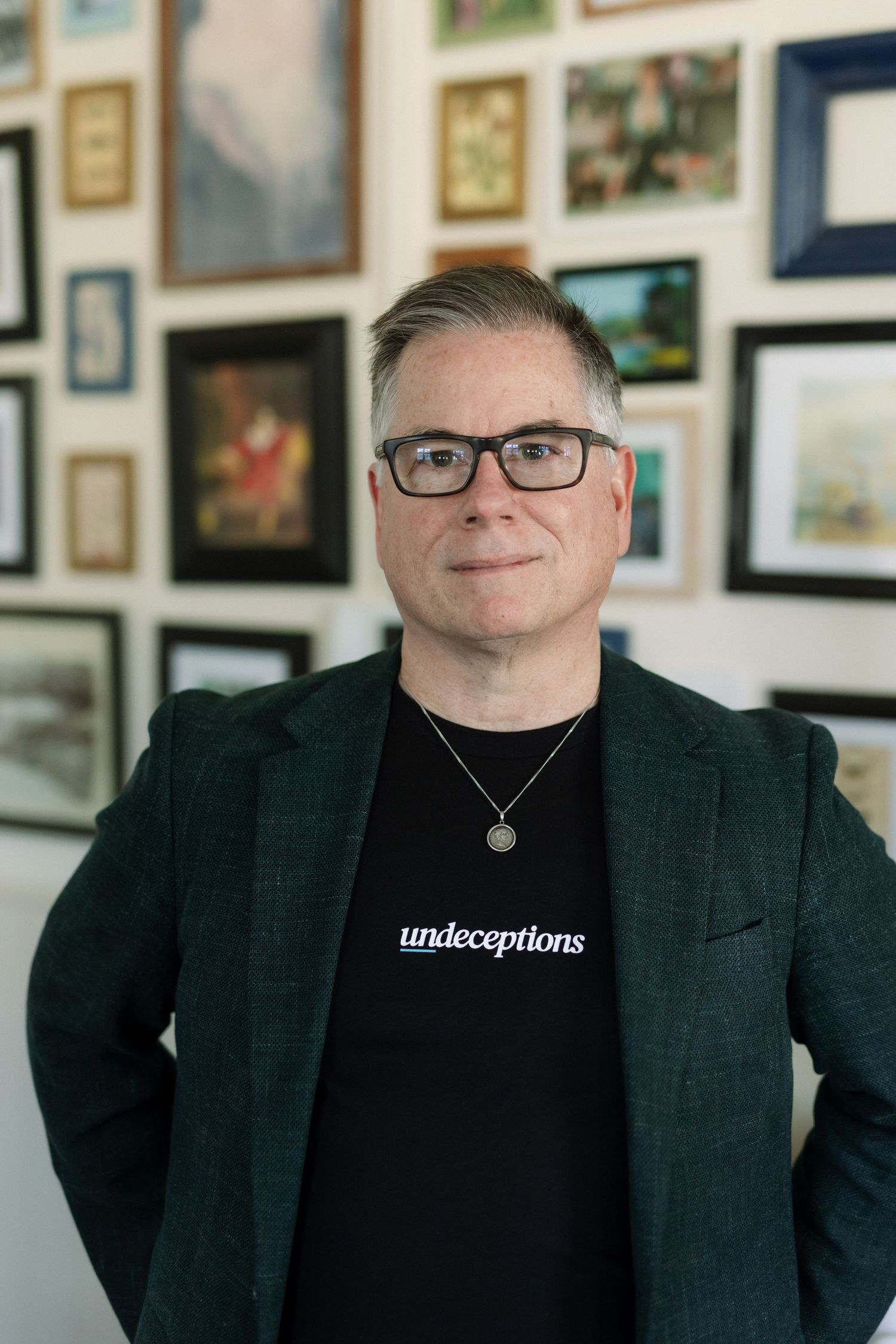 Man in dark blazer and Undeceptions t-shirt poses in front of gallery wall of framed artwork.