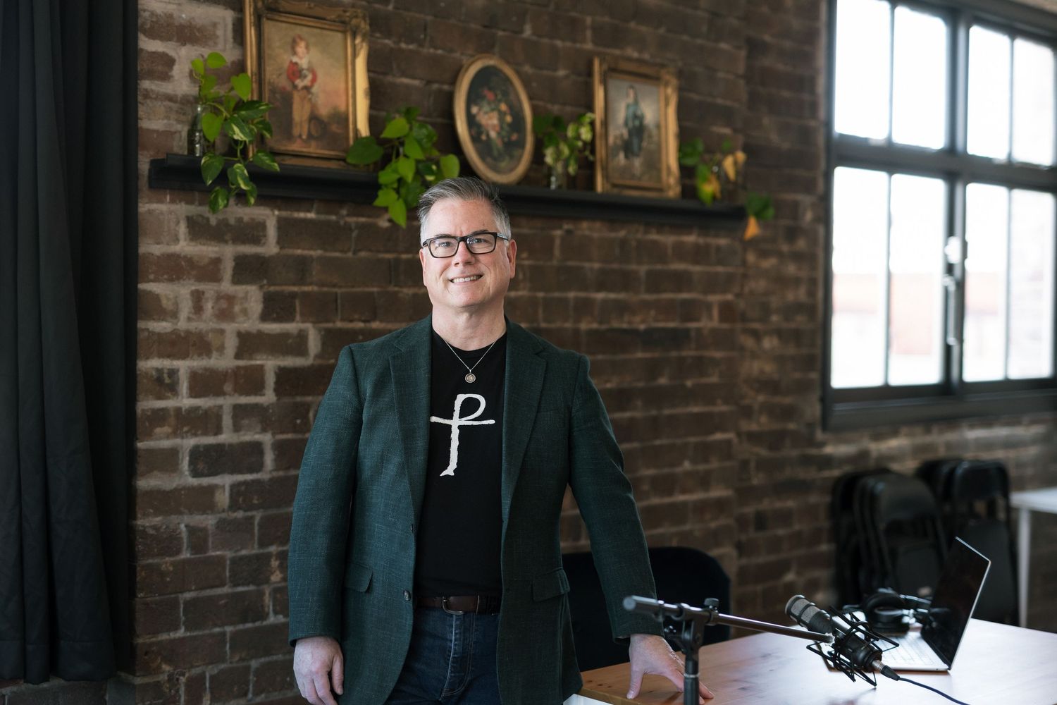 Person in green blazer and black t-shirt standing in a brick-walled office with framed artwork and plants.
