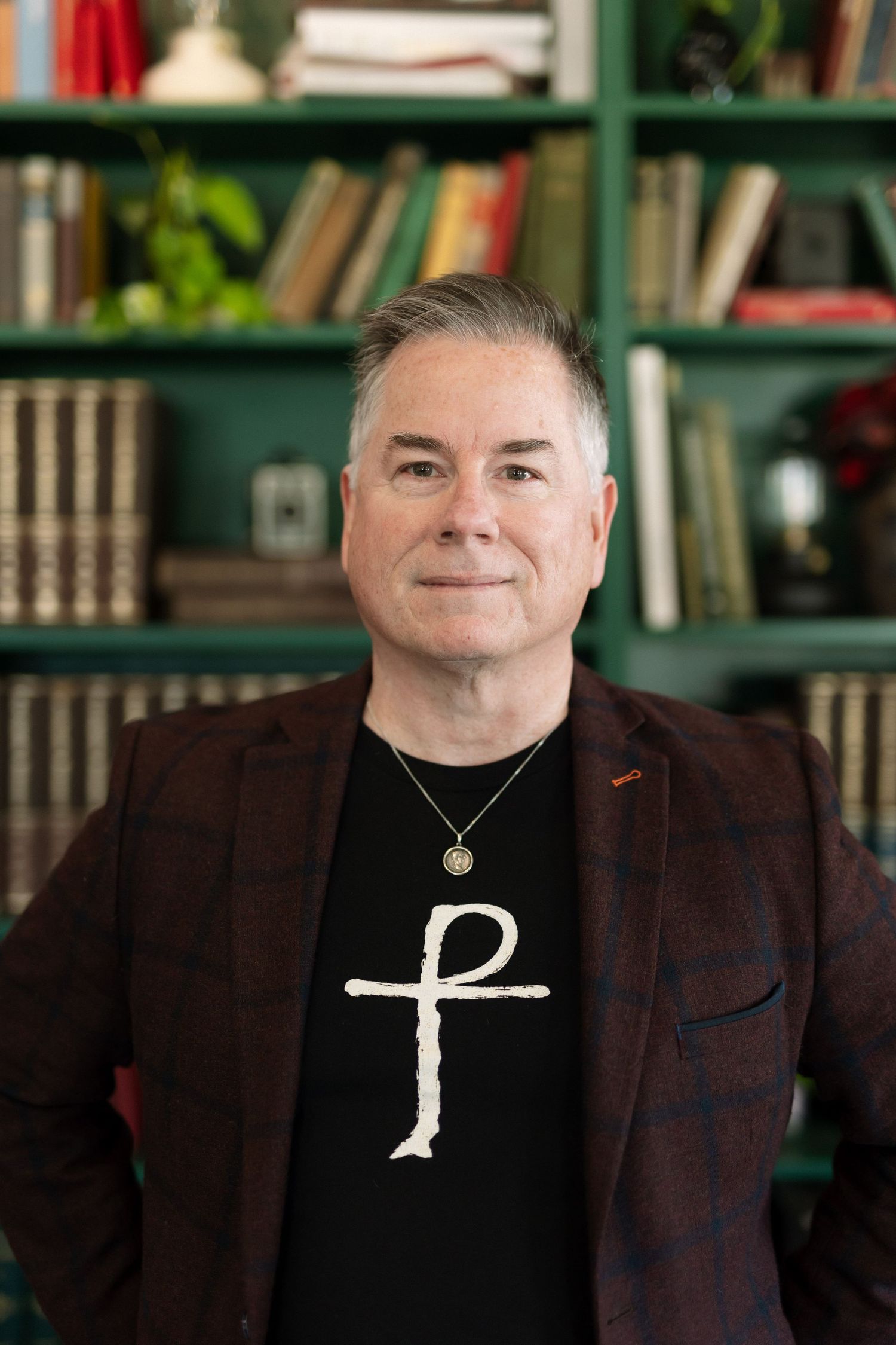 Man in plaid blazer and cross t-shirt smiling in front of green bookshelf filled with books.