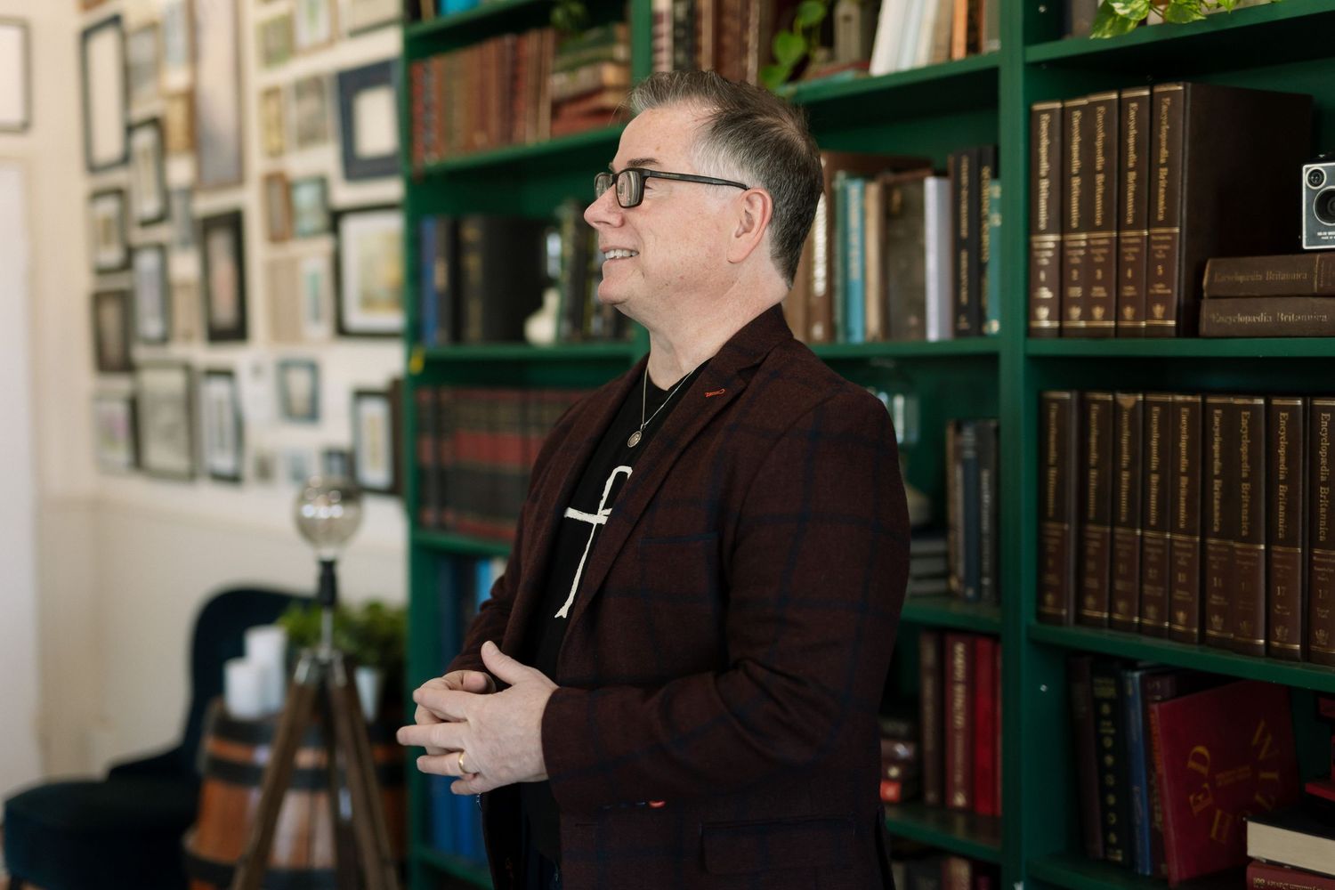 Man in dark blazer with glasses stands before green bookshelf filled with leather-bound books in a cozy library setting.
