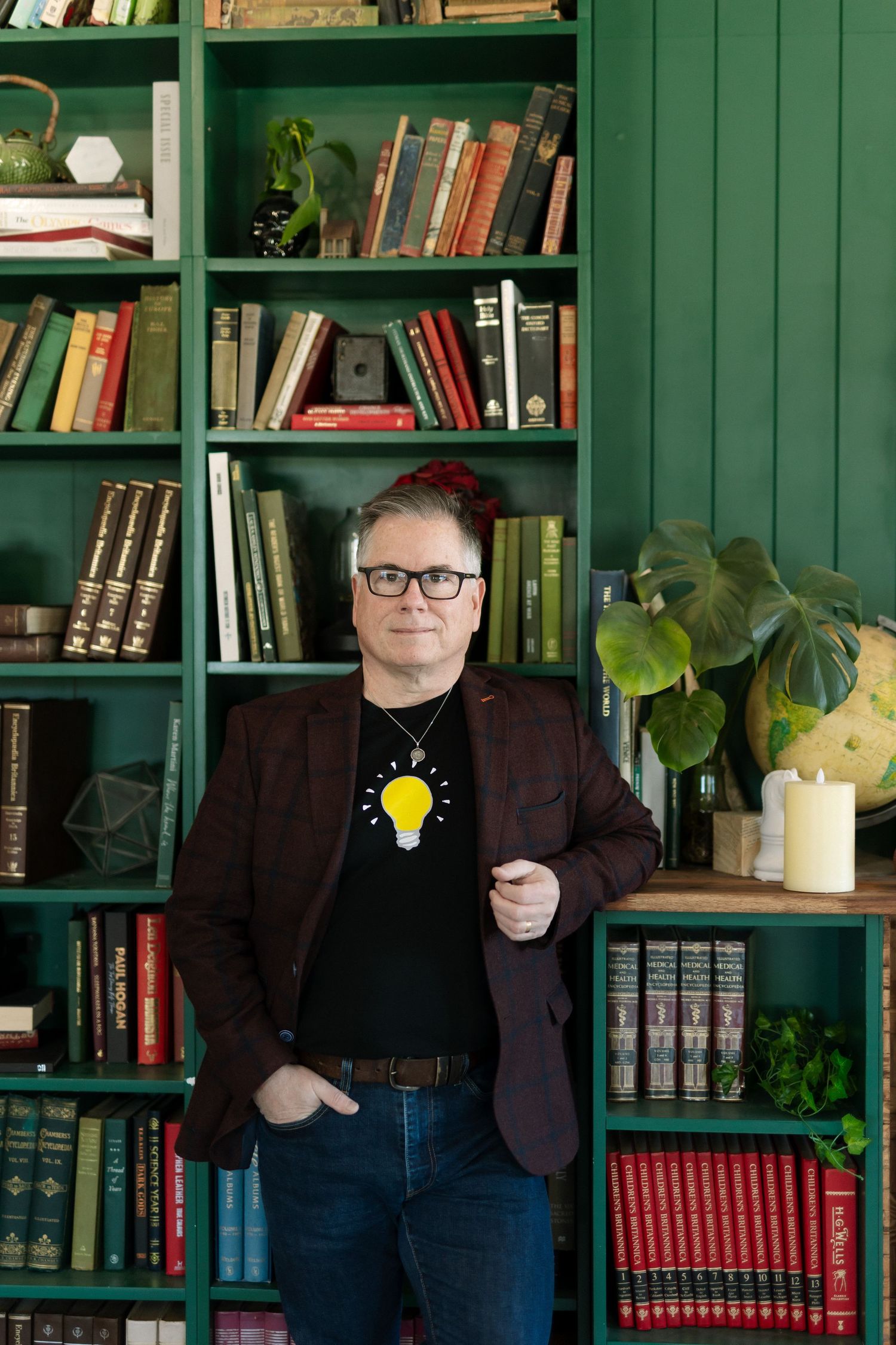 Man in blazer and lightbulb tee stands before green shelves filled with books and plants.