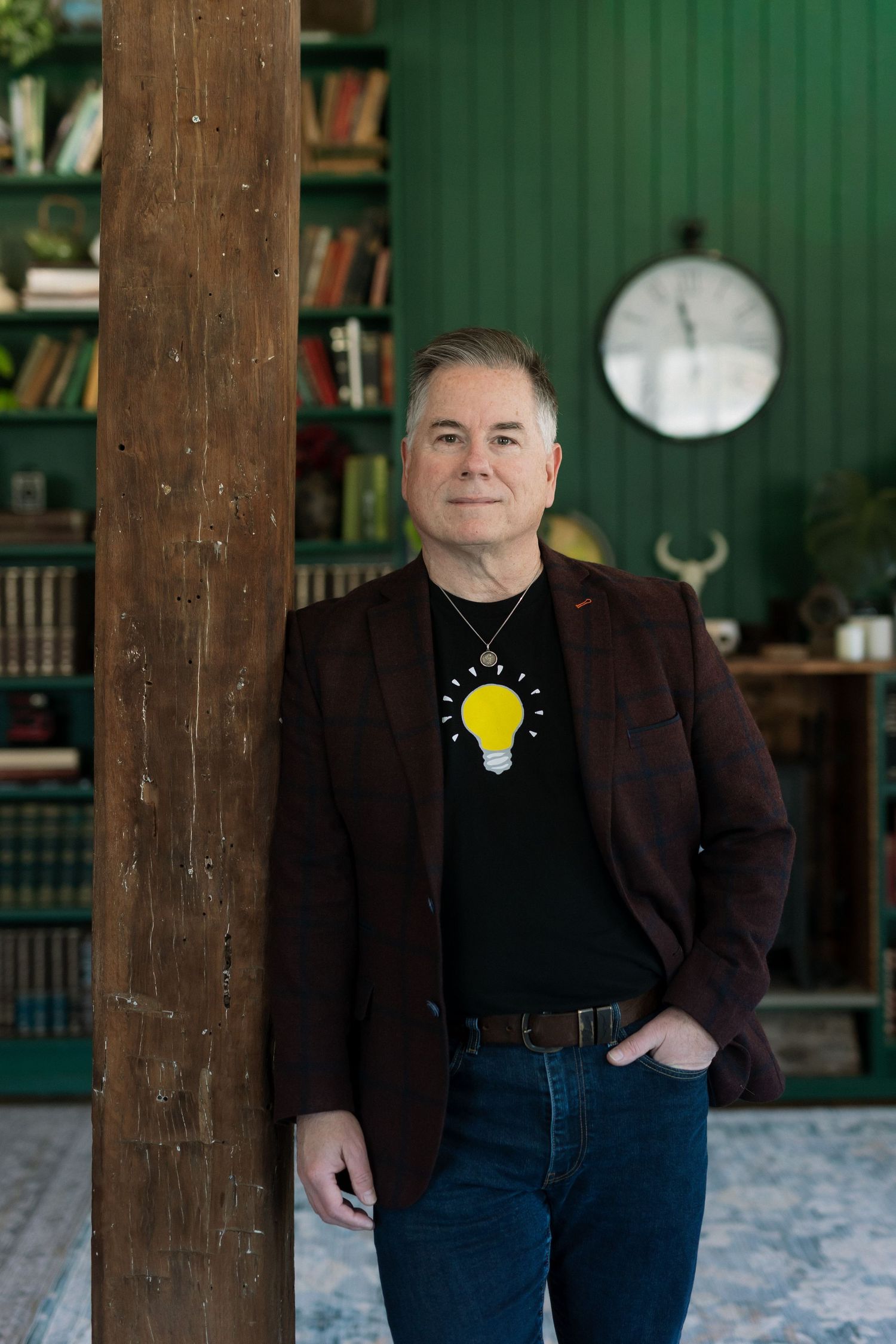 Man in dark blazer and light bulb graphic tee leans against wooden pillar in green library room.